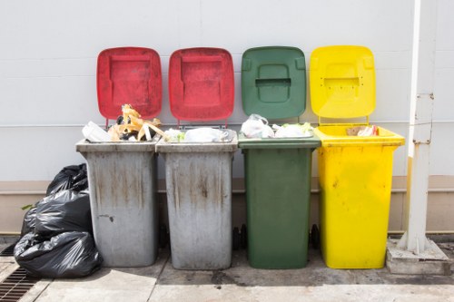 The image shows four large wheelie bins aligned side by side on a concrete surface in front of a white wall. The first bin on the left is grey with its red lid open, revealing mixed waste including paper and plastic. The second bin, also grey with its lid open, contains similar waste materials. The third bin is green with a closed lid, and the fourth bin on the right is bright yellow, also with a closed lid. To the left of the bins, there are black plastic rubbish bags piled on the ground, partially resting on a metal grate. The scene appears to be part of a waste disposal area, possibly within a residential or commercial property, with minimal surrounding elements visible. This setup is typical for rubbish removal services like those provided by Kilburn House Clearance, helping to manage household or business waste efficiently in the Kilburn or nearby areas of north west London.