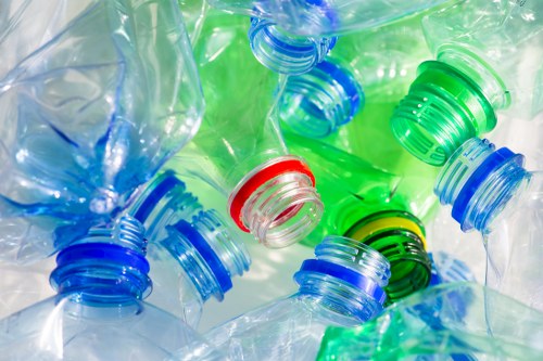 Close-up image of numerous transparent plastic bottles with coloured caps, including red, blue, green, and black, piled together with some overlapping and tilted in various directions. The bottles appear to be empty, with visible ridges on the necks and textured caps. The background is blurred, and the lighting highlights the glossy surfaces and vibrant colours of the caps and bottles. This assortment of plastic bottles, shown at close range, reflects common household or commercial waste typically handled during rubbish removal services in areas like Kilburn, London. The image emphasizes the importance of proper disposal and recycling of plastic waste, consistent with services provided by Kilburn House Clearance for domestic or commercial rubbish collection and clearance in the local vicinity.