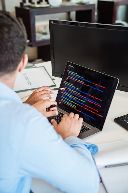 A person with short dark hair, wearing a light blue shirt, is seated at a white desk using a laptop with a black keyboard. The laptop screen displays colorful lines of code, including blue, red, yello