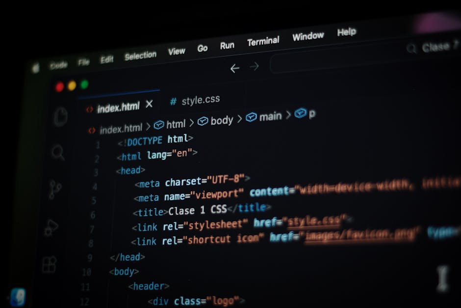 A person with short dark hair, wearing a light blue shirt, is seated at a white desk using a laptop with a black keyboard. The laptop screen displays colorful lines of code, including blue, red, yello